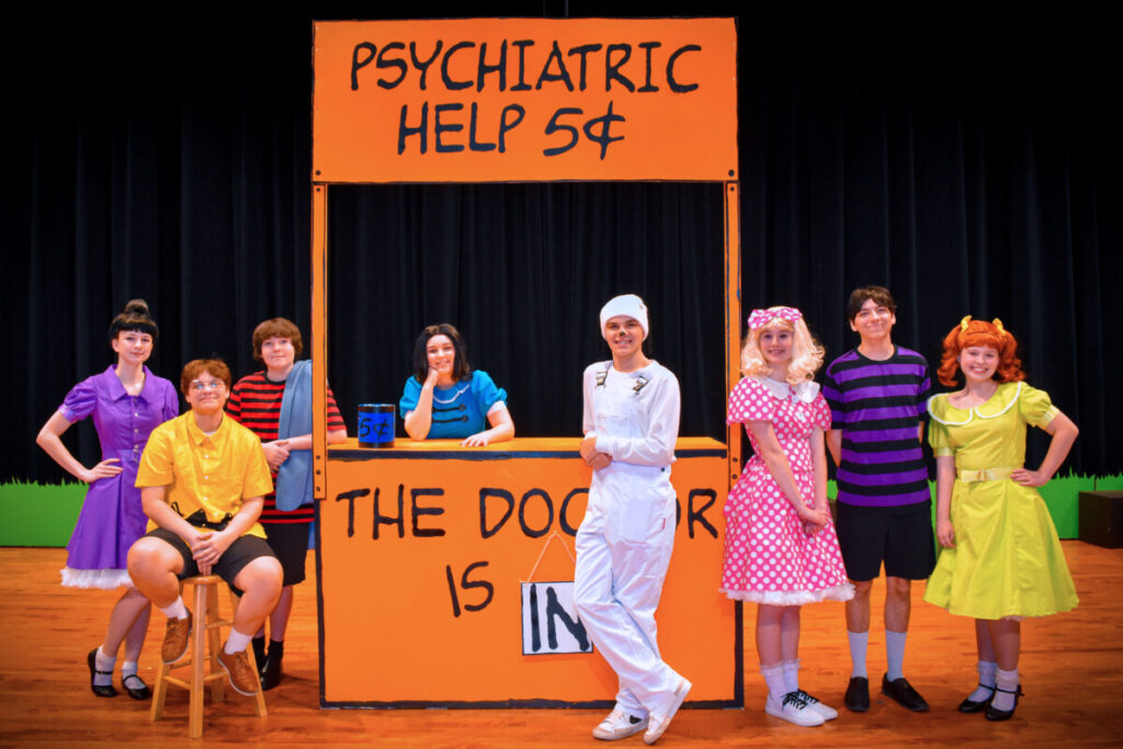 • The cast of You're a Good Man, Charlie Brown gathers around Lucy's iconic help booth, brought to life by Ella Games, capturing a classic moment from the Peanuts world.