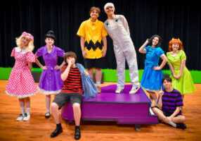 The cast of You're a Good Man, Charlie Brown gathers around Schroeder’s piano.